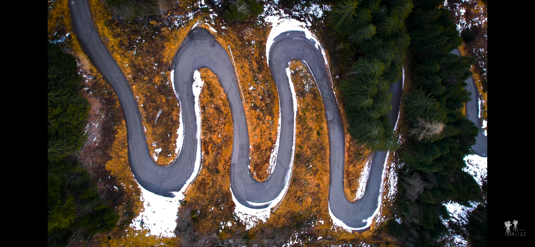 Zdjęcie z drona przedstawiające krętą asfaltową drogę przez las i łąki z resztkami śniegu.