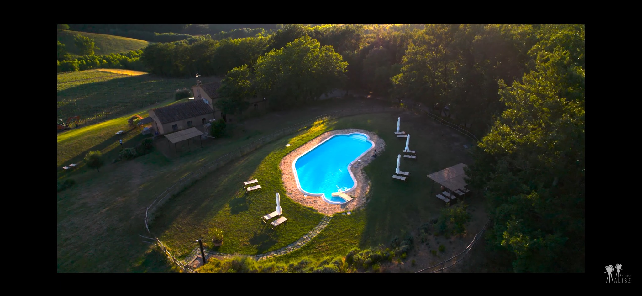 Panoramiczny widok z lotu ptaka na urokliwy basen o nieregularnym kształcie, otoczony zielonym trawnikiem, leżakami i parasolami, w scenerii toskańskiej willi z czerwonym dachem na wzgórzu...
