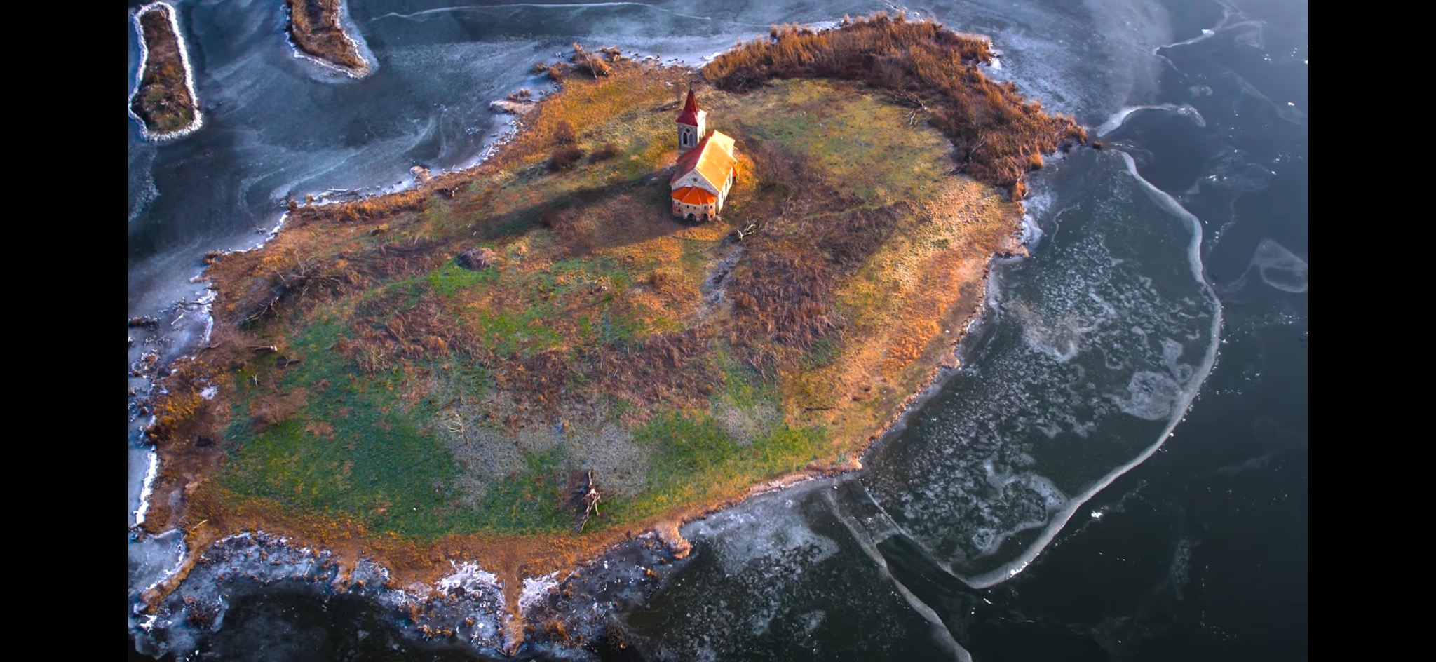 Widok z lotu ptaka na małą wyspę z kościołem, otoczoną częściowo zamarzniętym jeziorem.