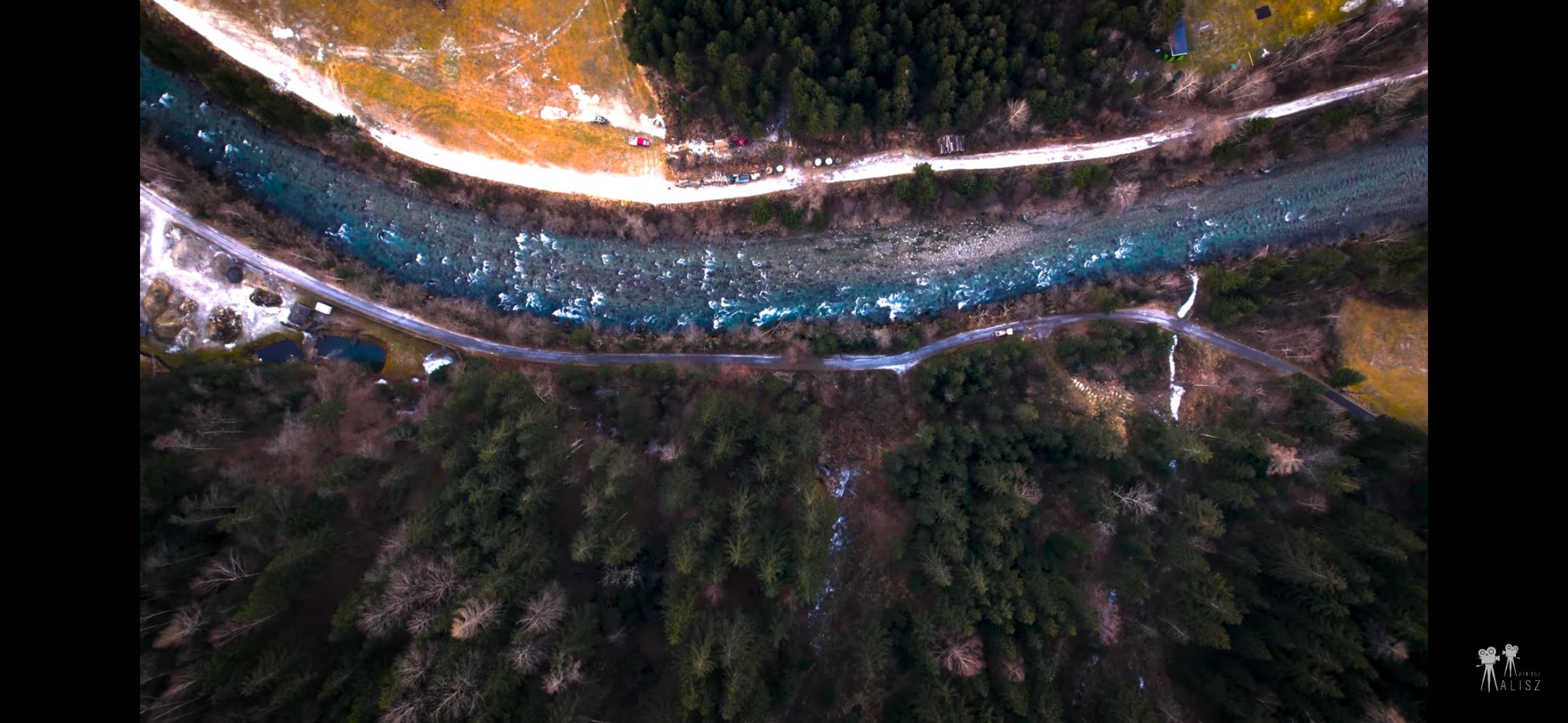 Panoramiczny widok z drona na rzekę z krystaliczną wodą, otoczoną lasem i drogą, z widocznym logo firmy wideofilmowania w prawym dolnym rogu.