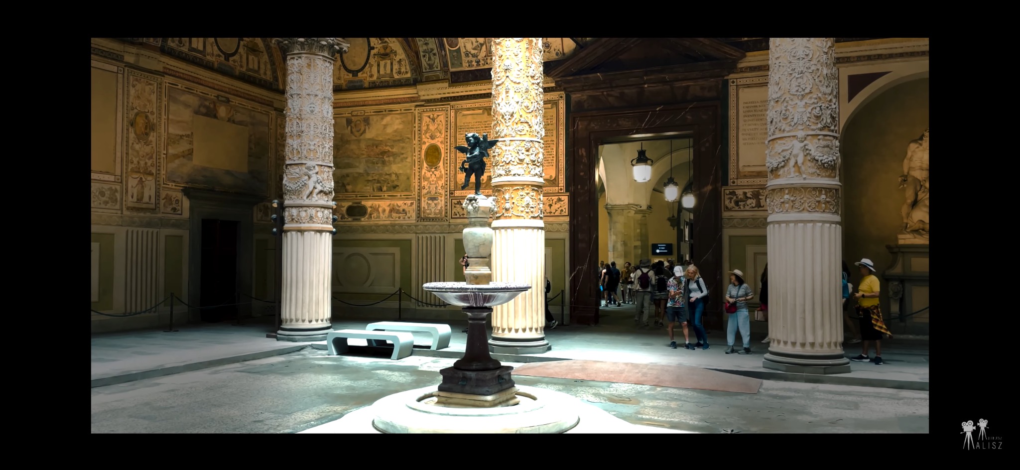 Wnętrze renesansowego pałacu z kolumnami zdobionymi płaskorzeźbami, fontanną pośrodku i turystami w tle, oświetlone naturalnym światłem wpadającym przez drzwi.