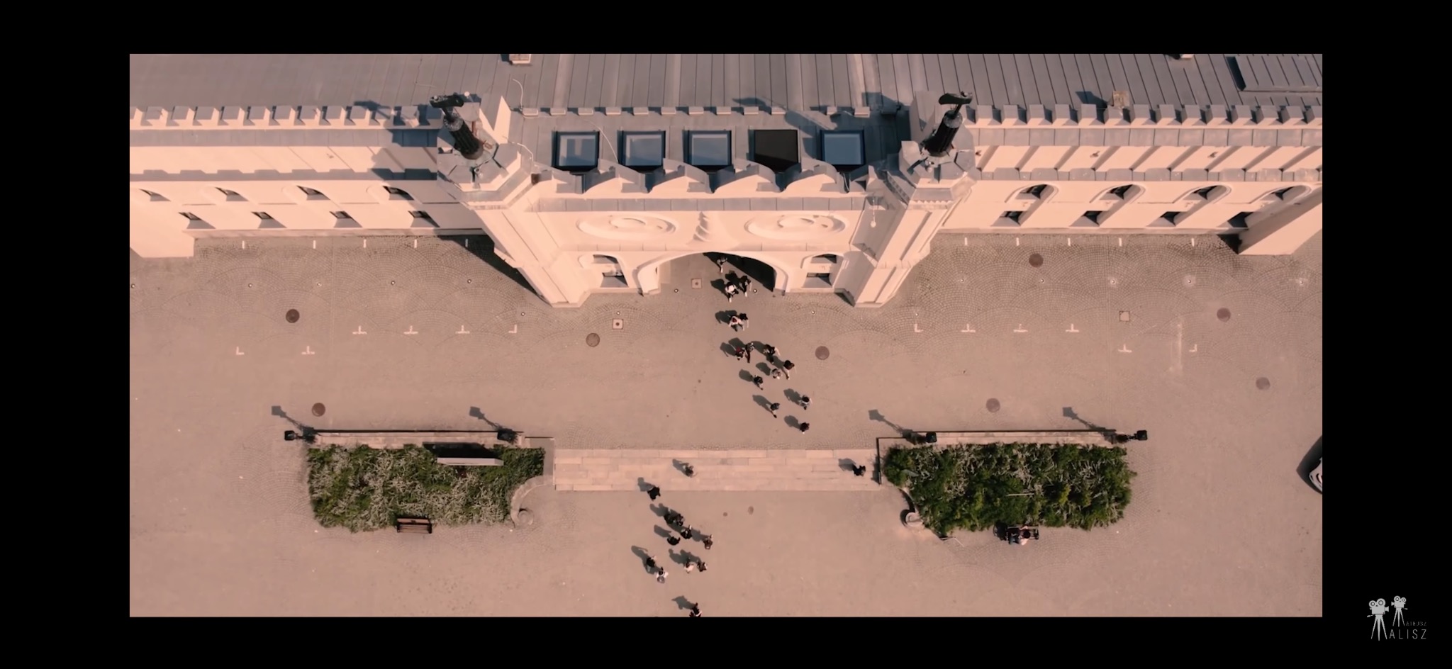 Szeroki kadr z lotu ptaka przedstawiający grupę ludzi wchodzących do budynku o architekturze zamkowej z jasnymi ścianami i szarym brukowanym placem.