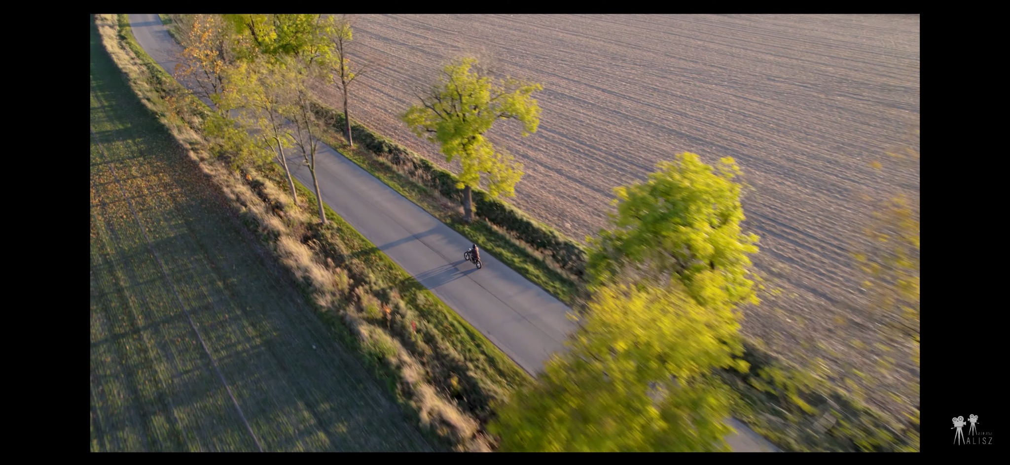 Ujęcie z drona przedstawiające motocyklistę jadącego asfaltową drogą, otoczoną polami i drzewami w jesiennej scenerii.