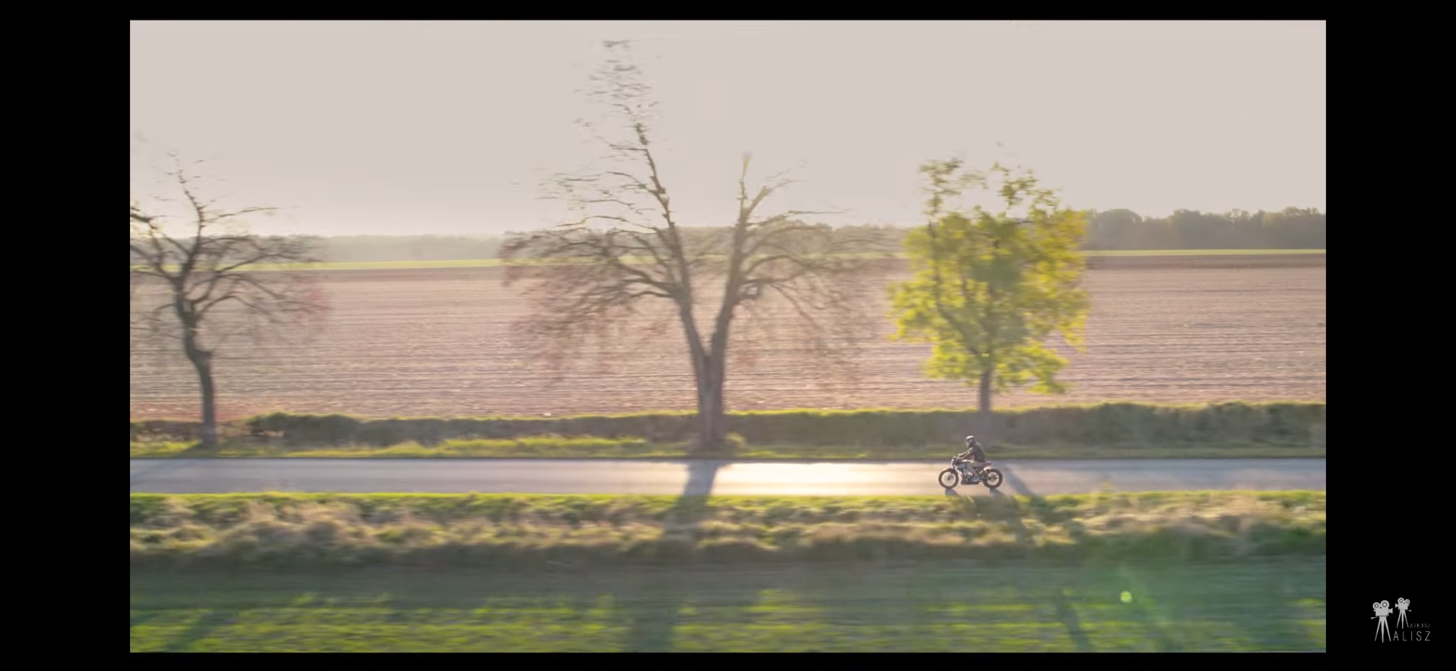 Ujęcie z drona: motocyklista na klasycznym motocyklu jedzie asfaltową drogą wzdłuż pola, w tle drzewa w jesiennej scenerii, ciepłe światło zachodzącego słońca.