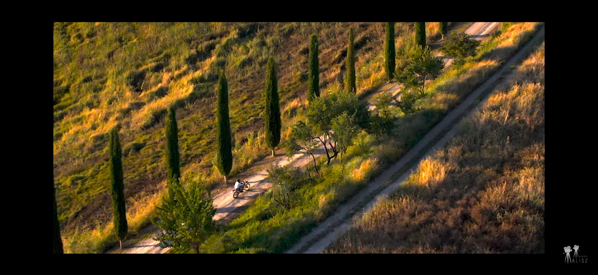 Motocyklista na szutrowej drodze, otoczony toskańskim krajobrazem z cyprysami, sfotografowany z lotu ptaka w ciepłym, popołudniowym świetle.