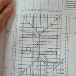 Fragment planu dachu z naniesionymi wymiarami i oznaczeniami elementów konstrukcyjnych, widoczny częściowo zakryty ręką i elementami biurowymi.