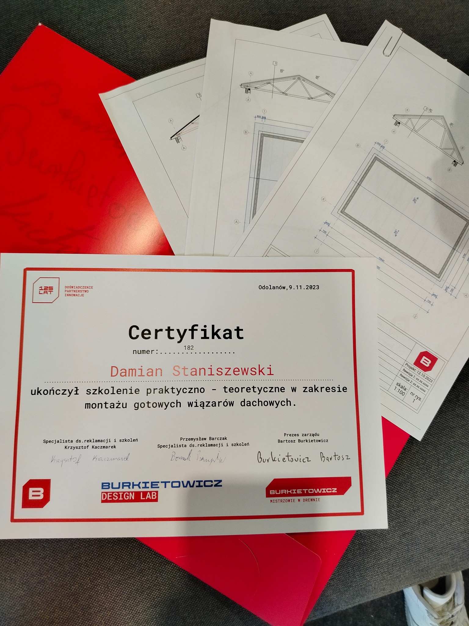 Certyfikat ukończenia szkolenia z montażu wiązarów dachowych Damiana Staniszewskiego, obok plany techniczne konstrukcji dachu i czerwona teczka.