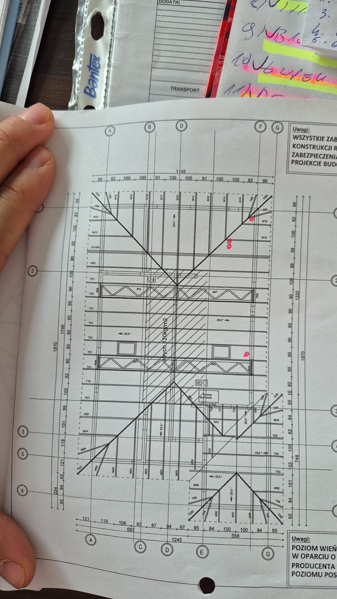 Fragment planu dachu z naniesionymi wymiarami i oznaczeniami elementów konstrukcyjnych, widoczny częściowo zakryty ręką i elementami biurowymi.