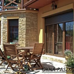 Zewnętrzne rolety w kolorze ciemnego brązu, zamontowane na oknach tarasowych budynku z elewacją z kamienia i żółtym tynkiem, widok z zewnątrz na drewniany zestaw mebli ogrodowych.