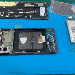 KMTechFix Kacper Wójcik - Rozłożony smartfon Samsung na niebieskiej macie serwisowej, widoczna bateria, wyświetlacz i wewnętrzne komponenty podczas naprawy.