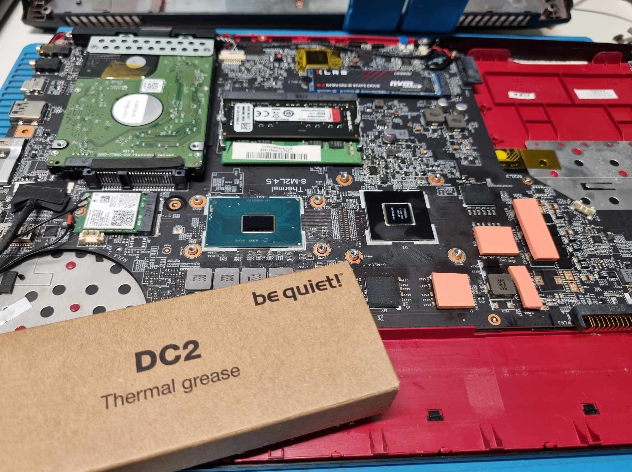 Otwarty laptop z widoczną płytą główną, dyskiem twardym, pamięcią RAM i kartą sieciową, obok pudełko pasty termoprzewodzącej 'be quiet! DC2 Thermal grease'.
