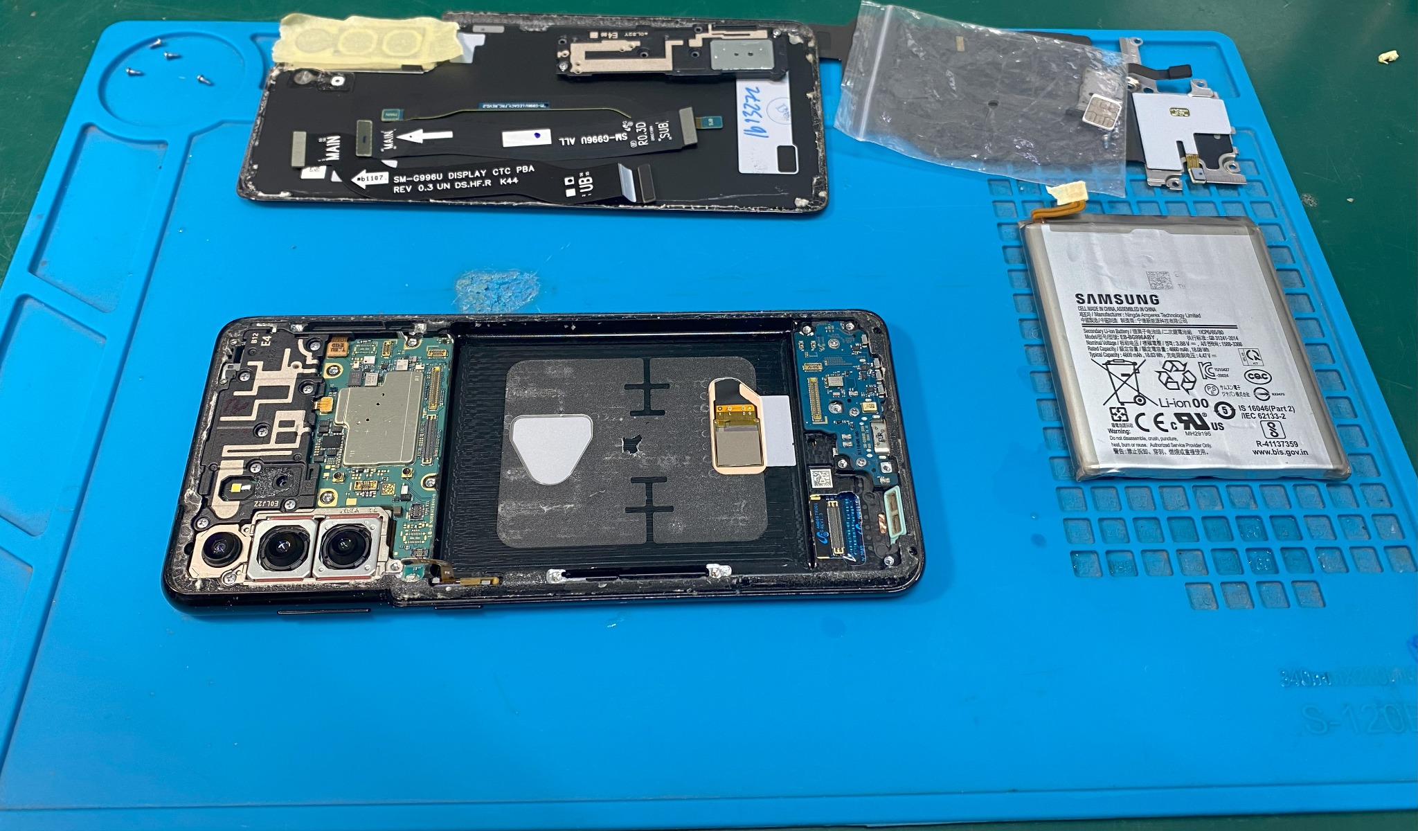 Rozłożony smartfon Samsung na niebieskiej macie serwisowej, widoczna bateria, wyświetlacz i wewnętrzne komponenty podczas naprawy.