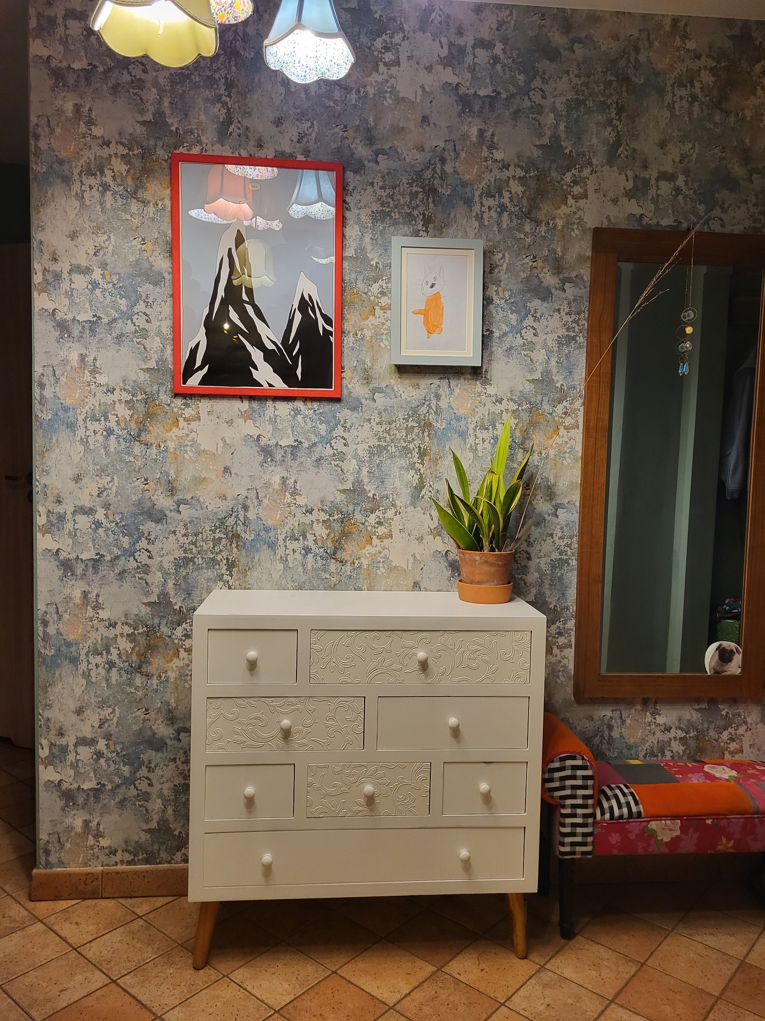 Biała komoda z dekoracyjnymi wzorami na szufladach, ustawiona na brązowej podłodze, na tle ściany z efektem postarzanego tynku, ozdobiona obrazami i lustrem.