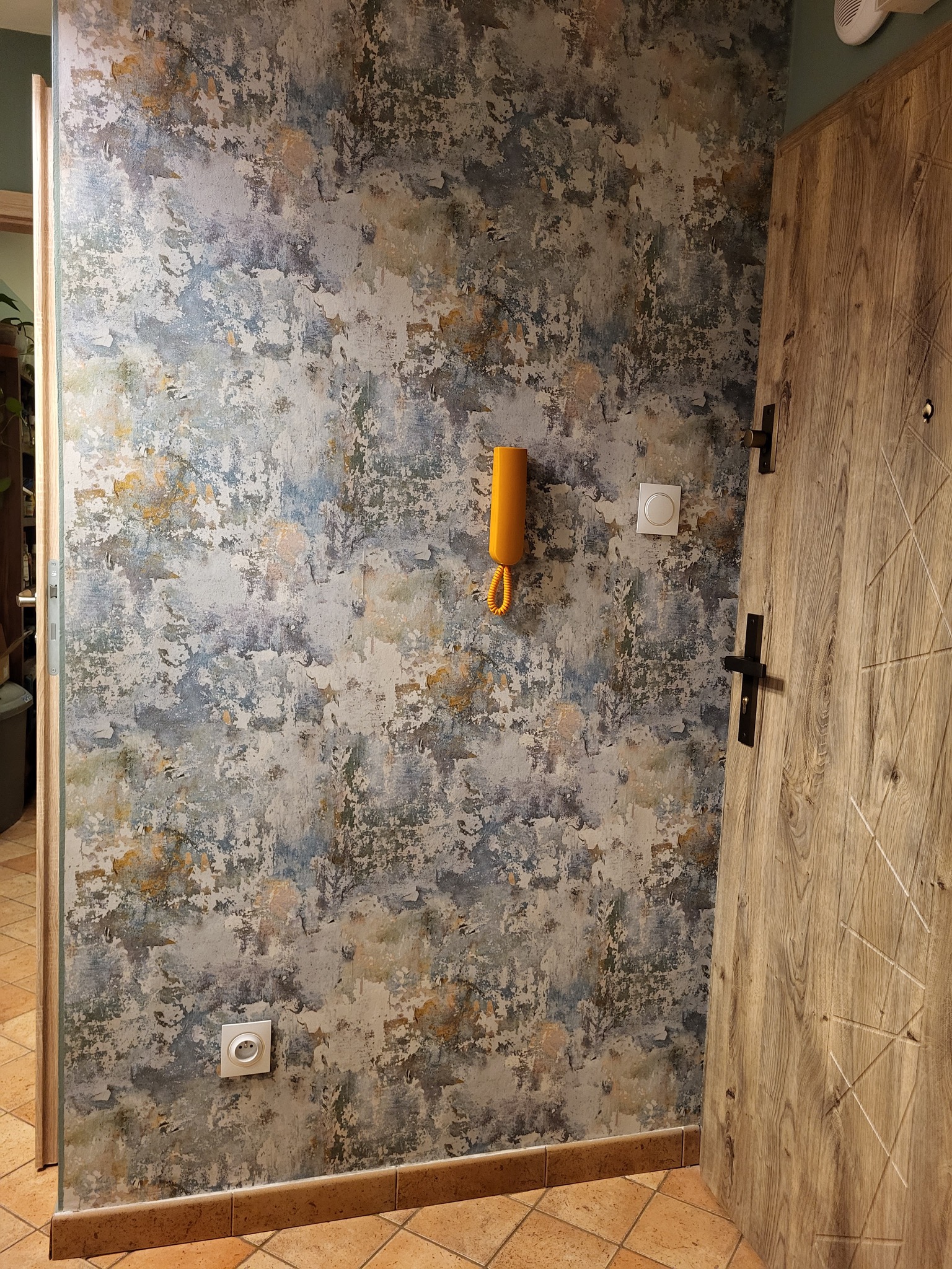 Ściana w mieszkaniu z dekoracyjną, wzorzystą tapetą w odcieniach szarości, błękitu i beżu, z zamontowanym żółtym domofonem i włącznikiem światła, obok drewnianych drzwi wejściowych z czarną klamką.