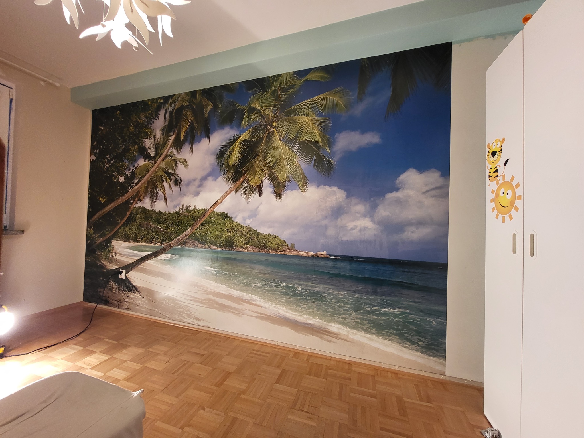 Fototapeta z motywem tropikalnej plaży z palmami na ścianie w pokoju, obok biała szafa z naklejkami, na podłodze parkiet.