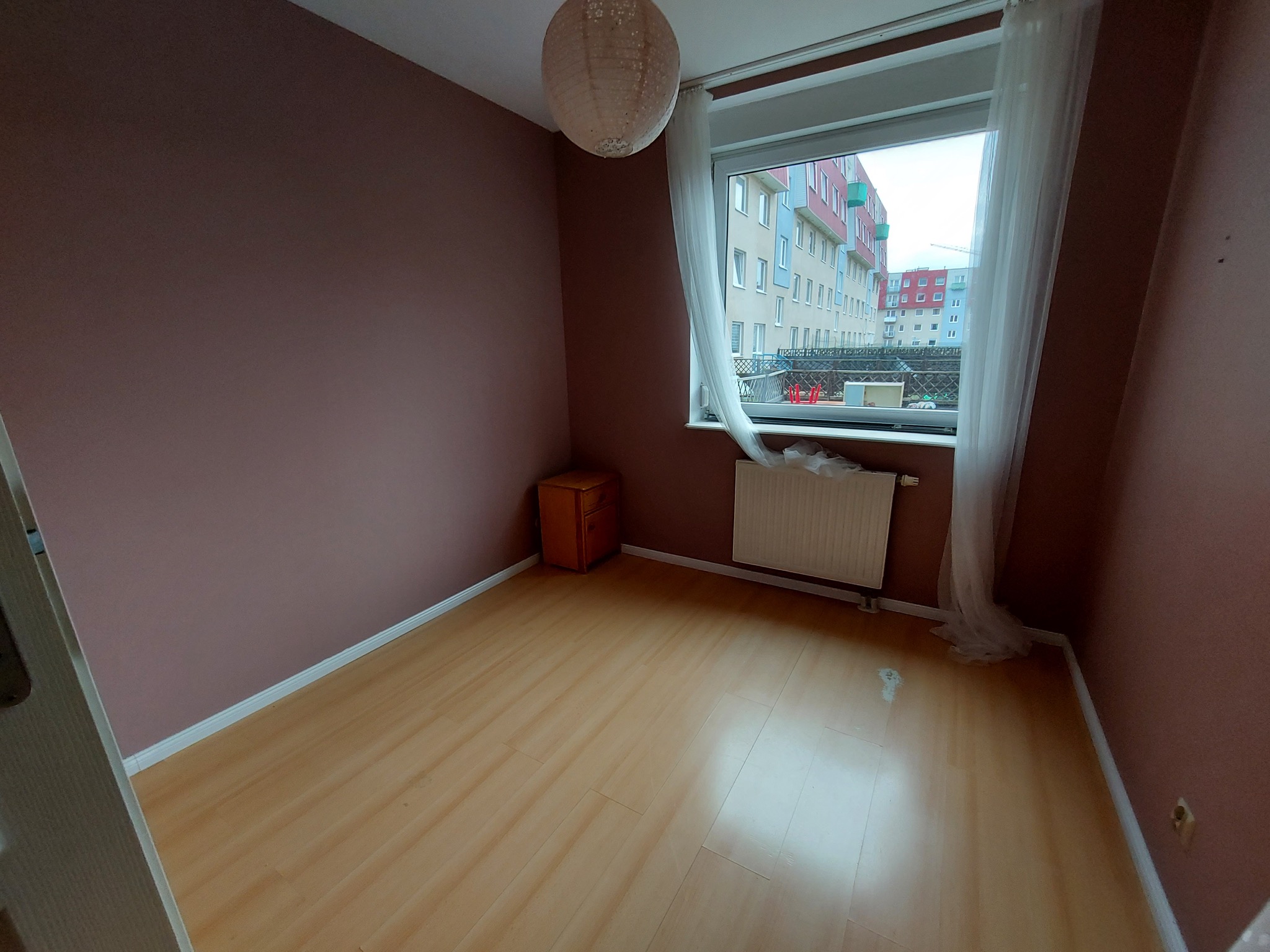 Pusty pokój z brązowymi ścianami, drewnianą podłogą, grzejnikiem pod oknem z białymi zasłonami i okrągłą lampą wiszącą. Widok na budynki mieszkalne za oknem.