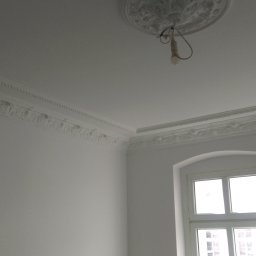 Rafał Zieliński - Białe wnętrze pokoju po remoncie, widoczne sztukaterie na suficie i ścianach, okno z białymi ramami oraz wisząca żarówka bez klosza.