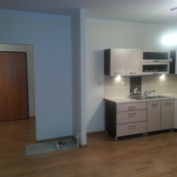 Rafał Zieliński - Jasna kuchnia z drewnopodobną podłogą po remoncie, widoczne szafki z oświetleniem podszafkowym, zlew, baterie i akcesoria kuchenne, na podłodze resztki silikonu i podkładu pod panele.