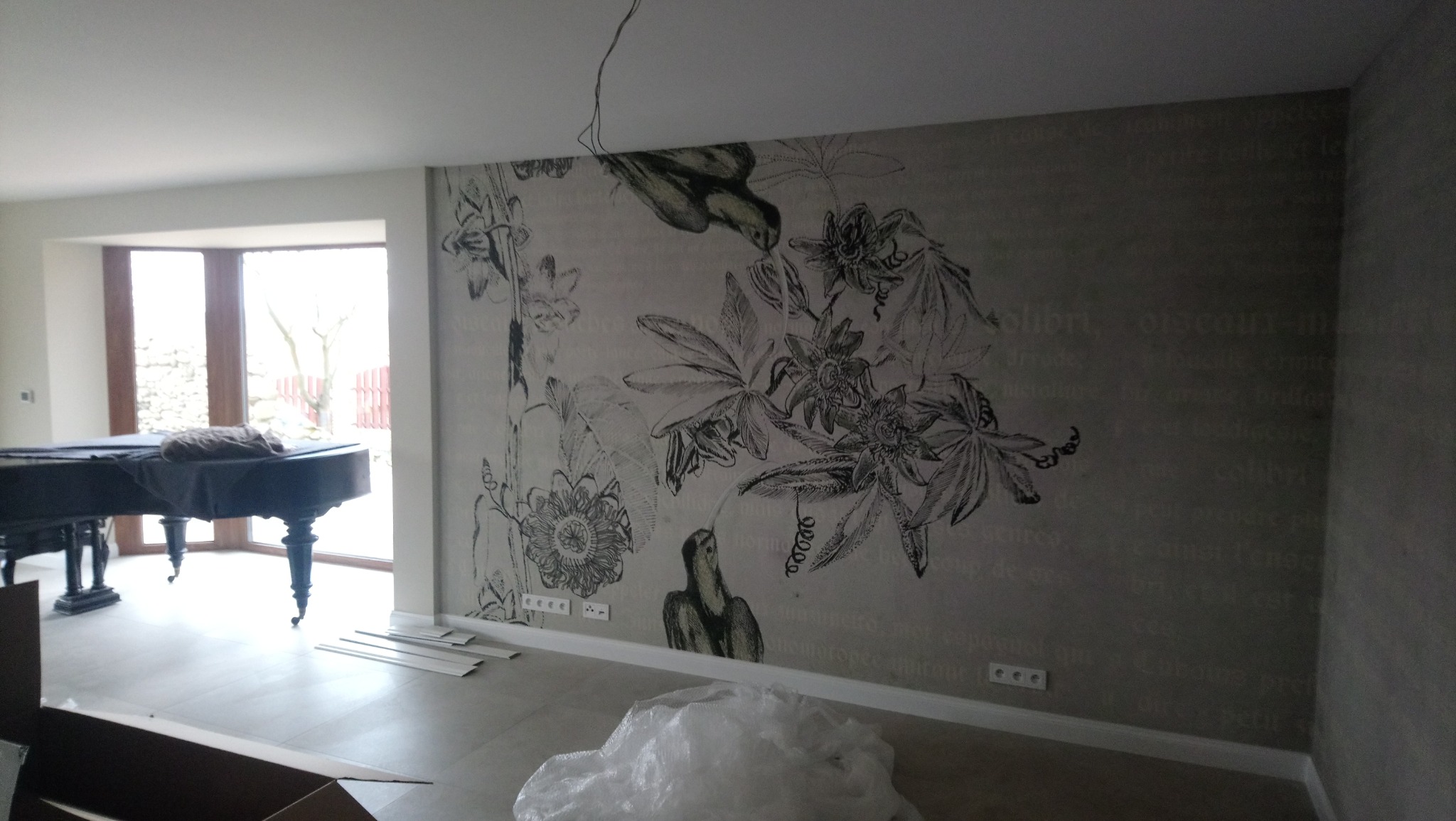 Pokój z pianinem i ścianą ozdobioną tapetą z motywem kwiatowym i wizerunkami ptaków, widoczne listwy przypodłogowe przygotowane do montażu.