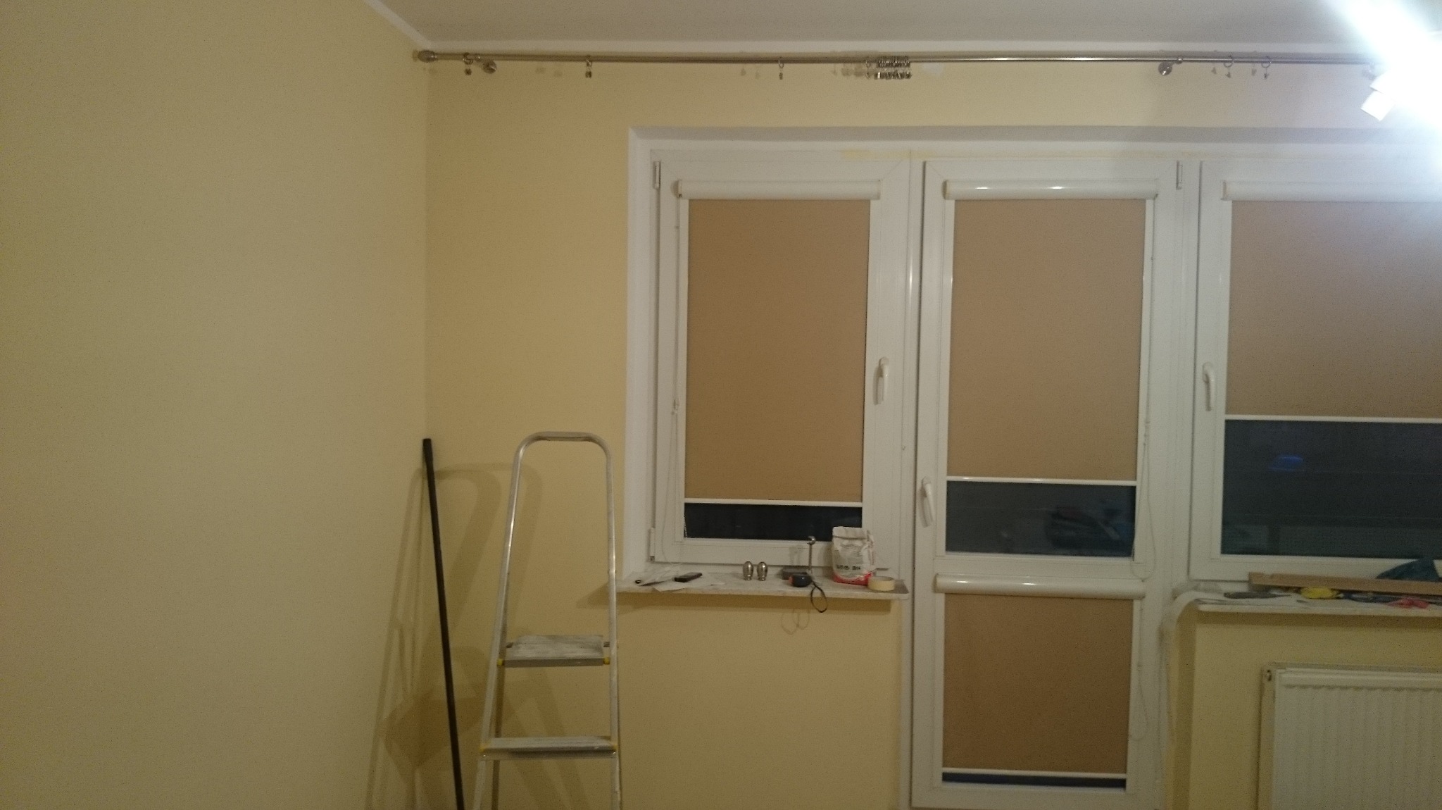 Żółta ściana w trakcie malowania, widoczna drabina aluminiowa, okno z roletami i przybory malarskie na parapecie, karnisz zamontowany nad oknem.