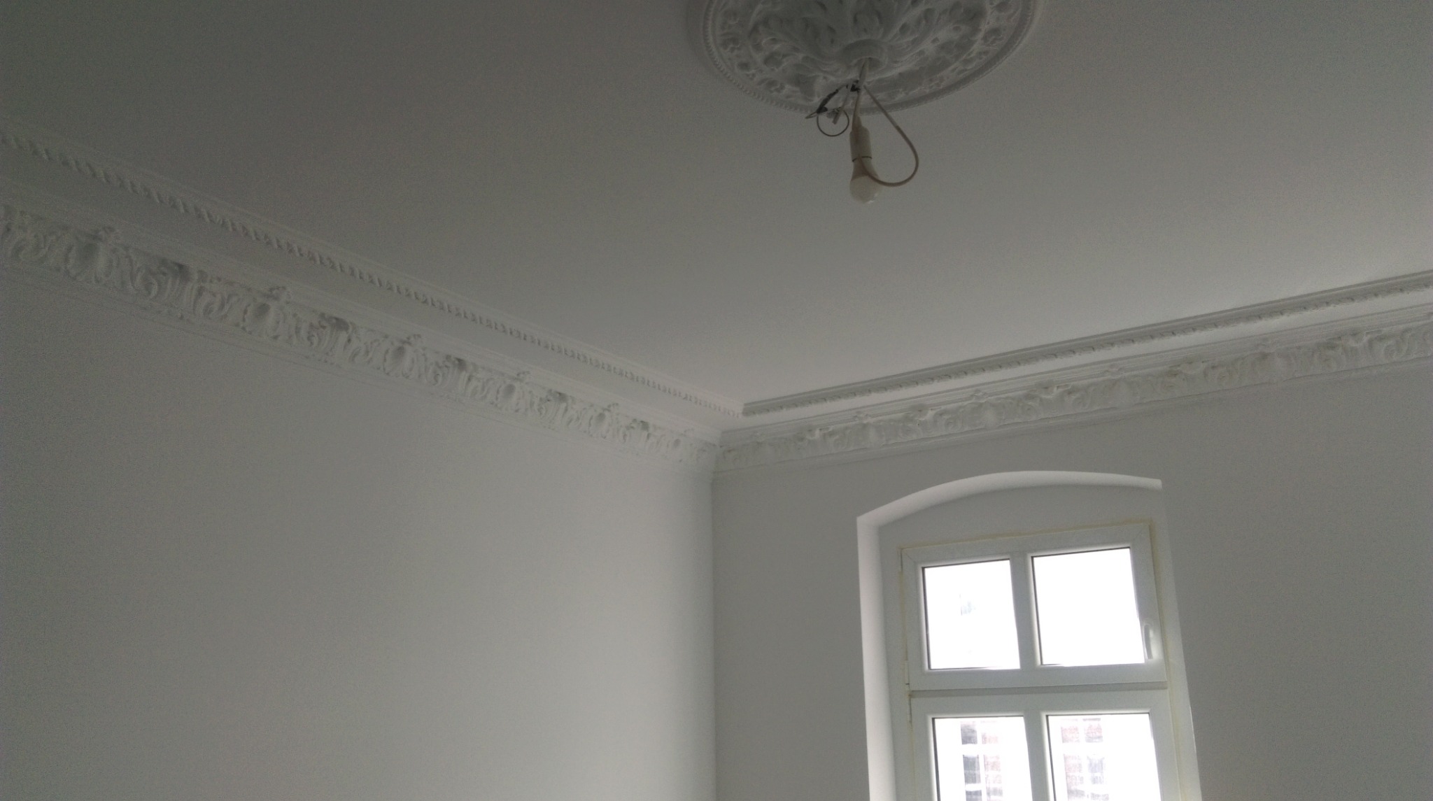 Białe wnętrze pokoju po remoncie, widoczne sztukaterie na suficie i ścianach, okno z białymi ramami oraz wisząca żarówka bez klosza.