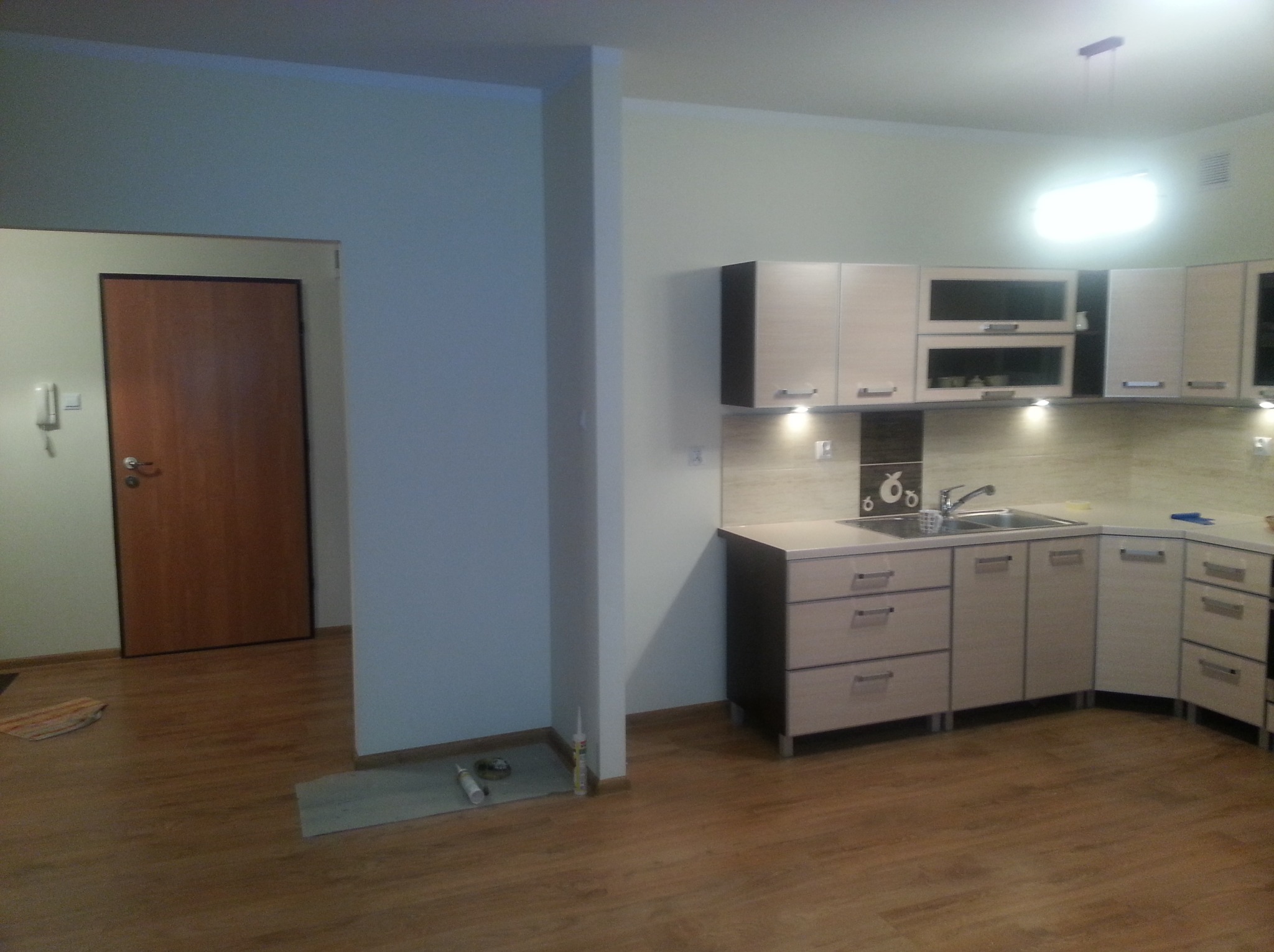 Jasna kuchnia z drewnopodobną podłogą po remoncie, widoczne szafki z oświetleniem podszafkowym, zlew, baterie i akcesoria kuchenne, na podłodze resztki silikonu i podkładu pod panele.