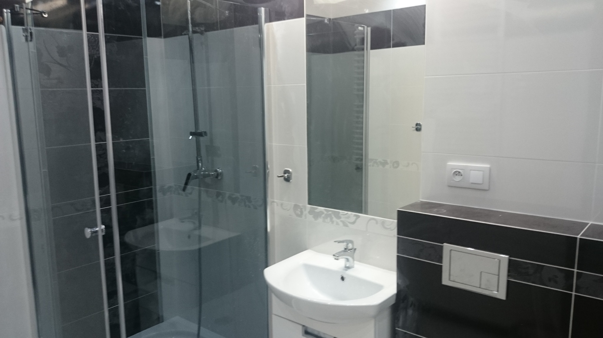 Wykończona łazienka z kabiną prysznicową z przesuwnymi drzwiami, białą umywalką z szafką, lustrem z dekoracyjnym motywem i czarno-białymi płytkami na ścianie.