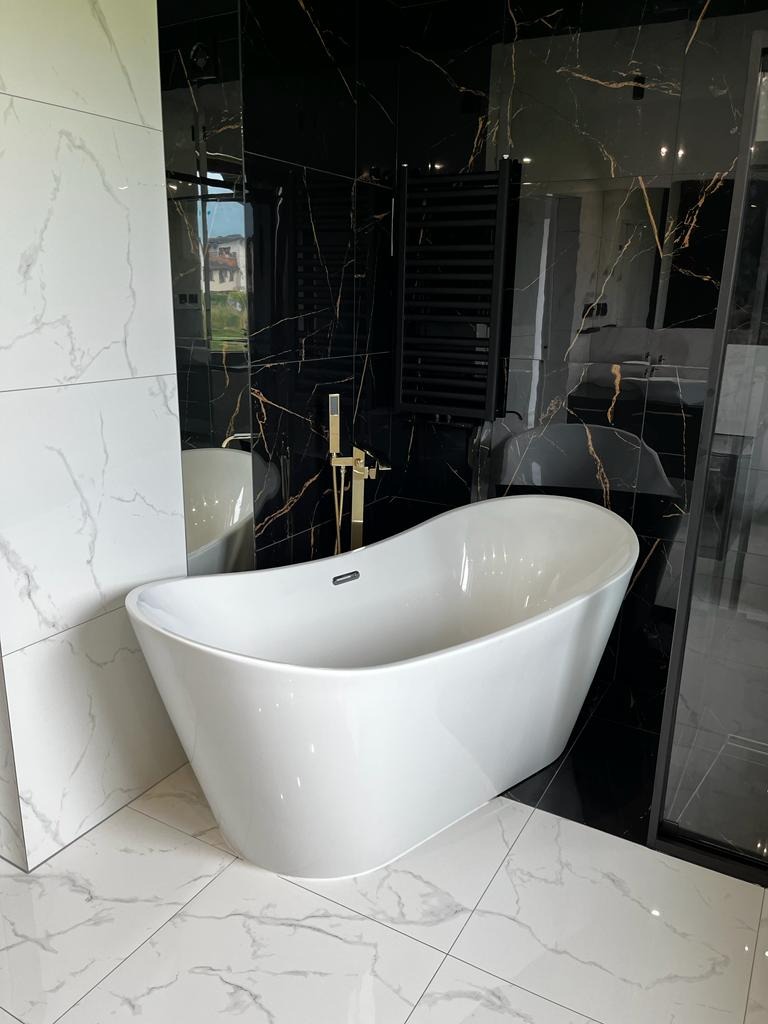 Nowoczesna łazienka z wolnostojącą białą wanną, złotą armaturą i czarnymi płytkami imitującymi marmur z złotymi żyłami, uzupełnona białymi płytkami imitującymi marmur na podłodze i ścianie.