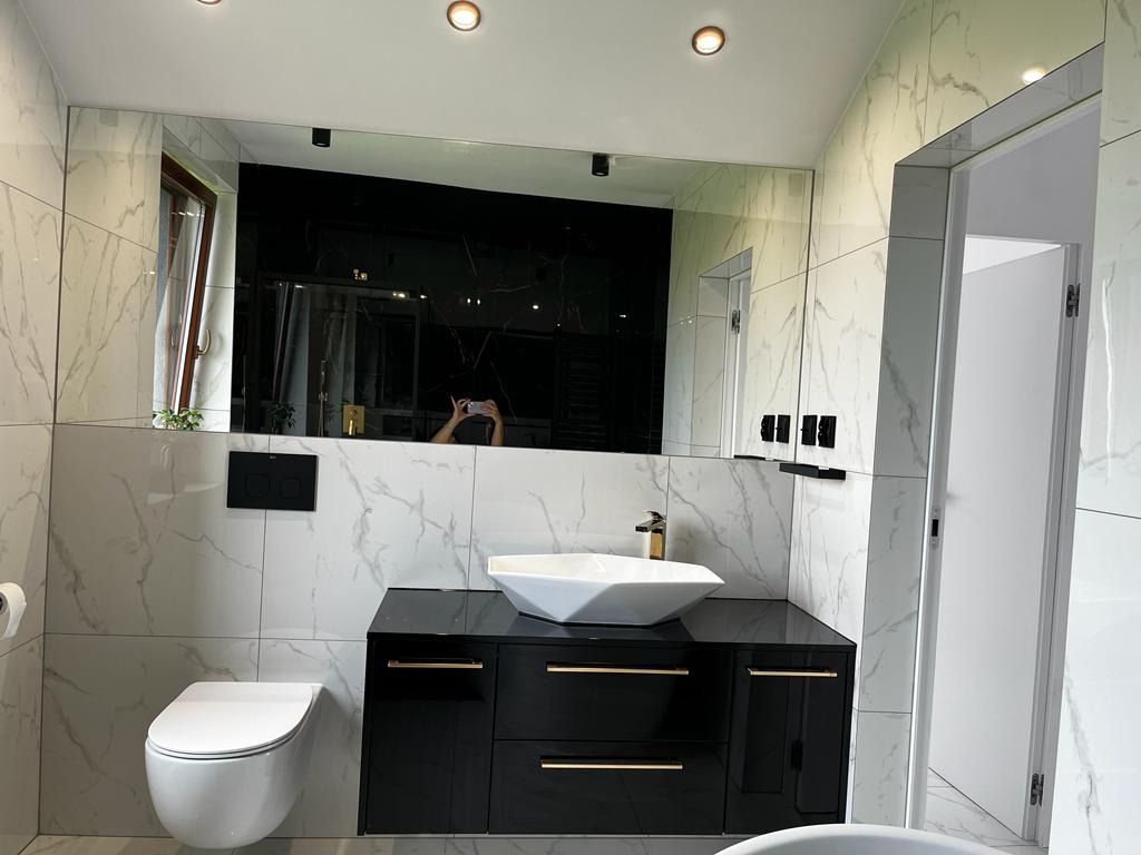 Nowoczesna łazienka z białymi marmurkowymi płytkami, czarną szafką z umywalką nablatową, złotą armaturą i dużym lustrem, w którym odbija się osoba robiąca zdjęcie.