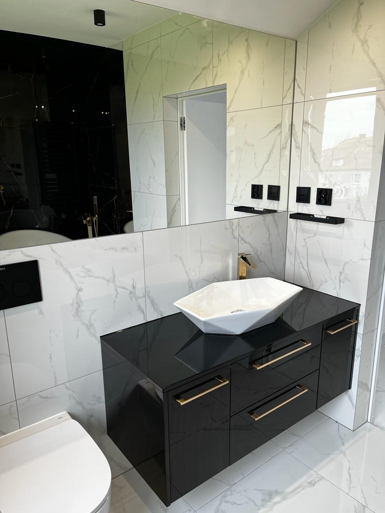 Nowoczesna łazienka z czarną, lakierowaną szafką pod umywalkę w kształcie diamentu, złotymi uchwytami i marmurowymi płytkami na ścianach i podłodze.
