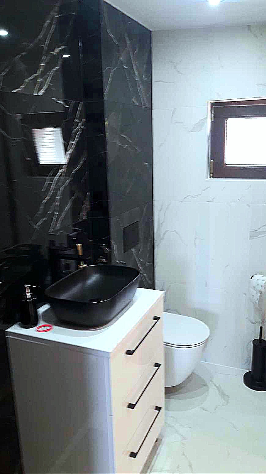 Nowoczesna łazienka z czarną umywalką nablatową, białymi płytkami imitującymi marmur i czarnymi płytkami z białymi żyłami na ścianie, biała szafka pod umywalką z trzema szufladami i czarnymi...