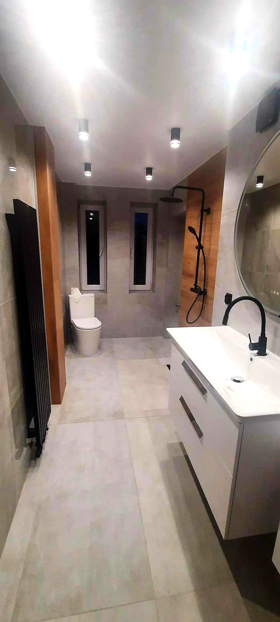 Nowoczesna łazienka z szarymi płytkami, czarną armaturą i grzejnikiem drabinkowym, oświetlenie punktowe, biała umywalka z szafką.