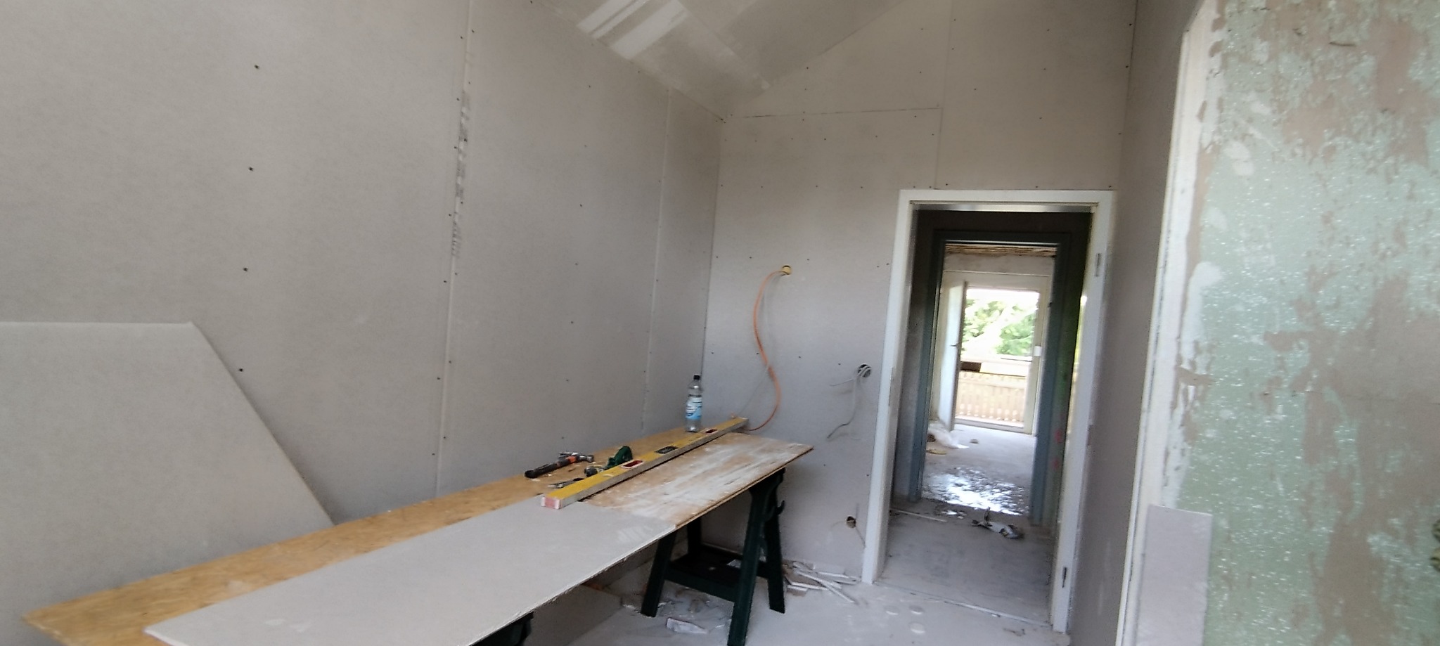 Wnętrze w trakcie remontu: ściany wyłożone płytami gipsowo-kartonowymi, widoczne narzędzia na stole roboczym, instalacja elektryczna wyprowadzona ze ściany, surowe wykończenie.