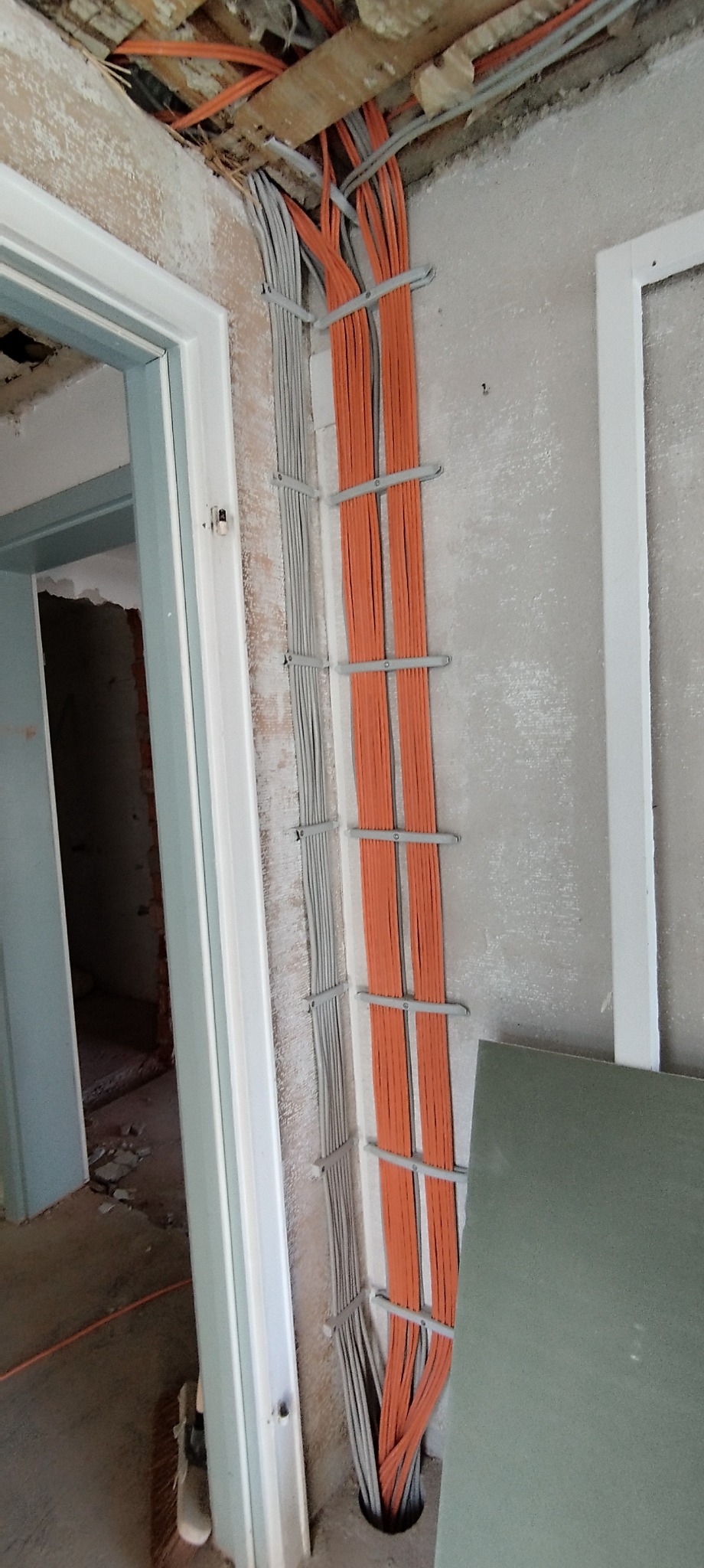 Pomarańczowe i szare przewody elektryczne ułożone równolegle w korytkach na ścianie w surowym wnętrzu, widoczne niedokończone ościeżnice drzwiowe.