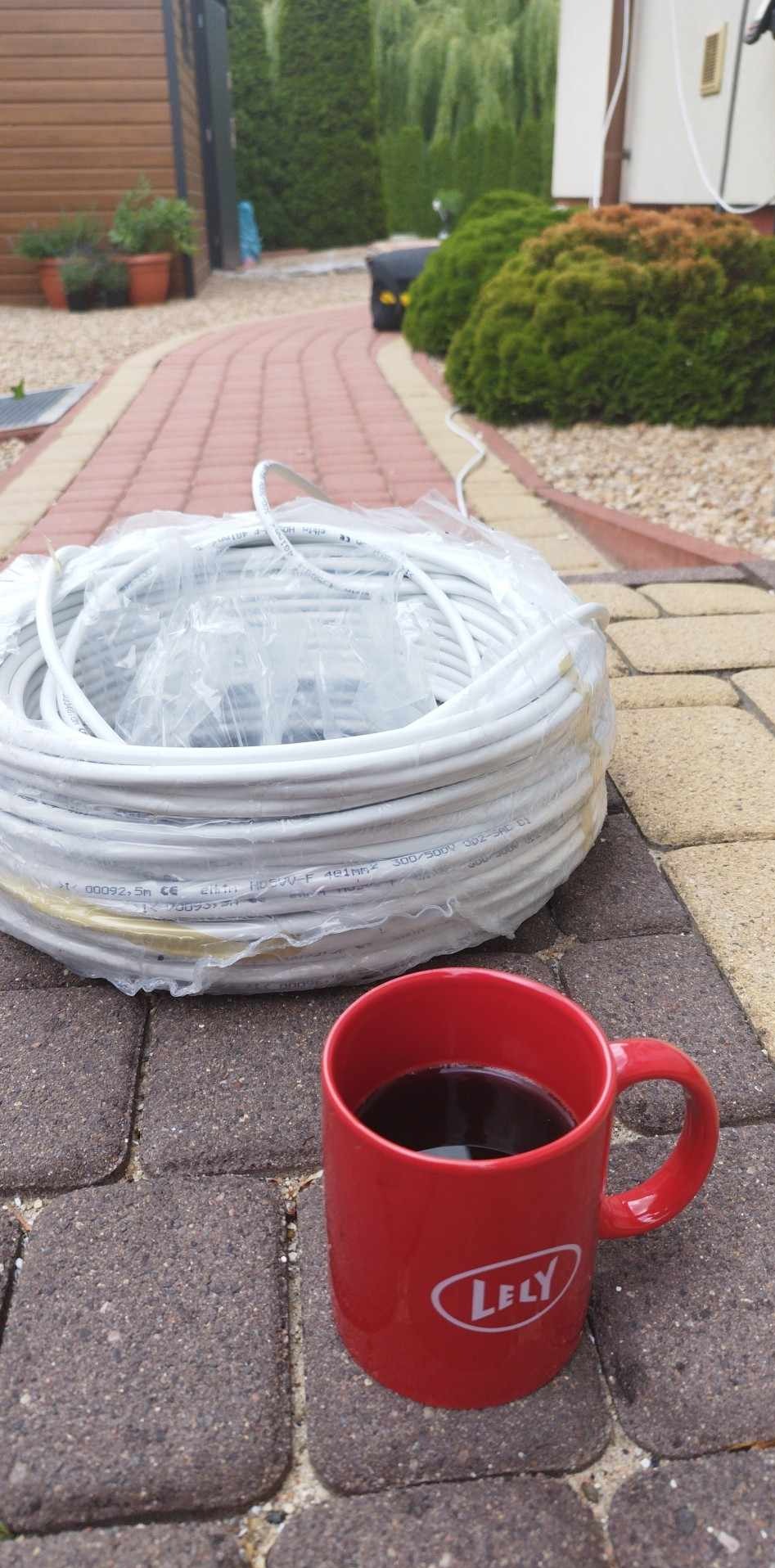 Zwój białego kabla elektrycznego w folii obok czerwonego kubka z napojem na brukowanej ścieżce, w tle zielone krzewy i budynek.
