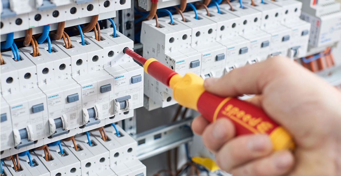 Ręka elektryka w trakcie pracy ze śrubokrętem przy zabezpieczeniach elektrycznych marki Siemens w rozdzielni, widoczne kable w kolorach niebieskim i brązowym.