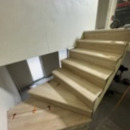 RIW remonty i wykończenia - Schody wyłożone jasnymi panelami drewnopodobnymi, widoczne pomarańczowe kliny dystansowe na jednym ze stopni, ściana w tle o nierównej fakturze, w prawym górnym rogu fragment odkurzacza przemysłowego.