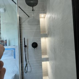 RIW remonty i wykończenia - Wykończona kabina prysznicowa z czarną armaturą, wnękami z oświetleniem i szarą płytką imitującą beton.