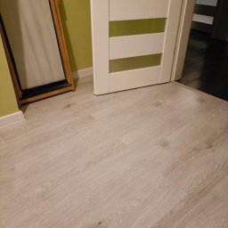 RIW remonty i wykończenia - Jasne panele podłogowe ułożone w nowym mieszkaniu, widoczna biała listwa przypodłogowa, częściowo widoczna biała, wewnętrzna stolarka drzwiowa.