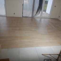 RIW remonty i wykończenia - Jasne panele podłogowe ułożone w nowym pomieszczeniu, widoczna lampa budowlana i zabezpieczone folią okna.