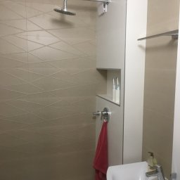 RIW remonty i wykończenia - Nowoczesna kabina prysznicowa z beżowymi płytkami o geometrycznym wzorze, chromowaną armaturą i wnęką z elektrycznymi szczoteczkami do zębów, uzupełniona o czerwoną wiszącą szmatkę i umywalkę...