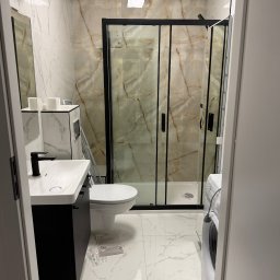 RIW remonty i wykończenia - Nowoczesna łazienka z kabiną prysznicową z czarnymi ramami, umywalką z czarną szafką, toaletą i pralką. Marmurowe płytki na ścianach i podłodze.