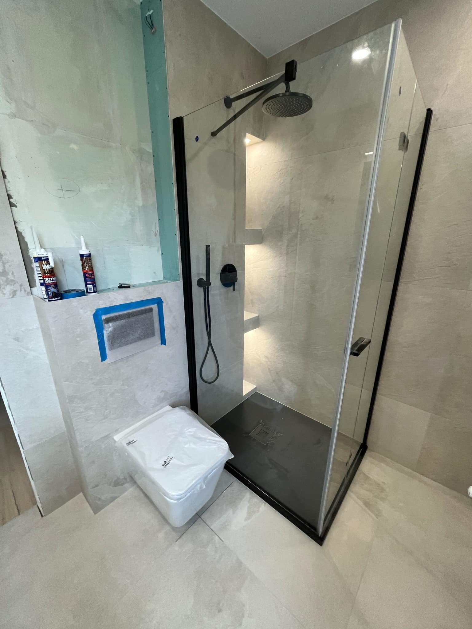 Remont łazienki: kabina prysznicowa z czarnymi detalami i deszczownicą, obok podwieszana toaleta zabezpieczona folią, ściany w trakcie wykończenia z widocznymi pojemnikami z silikonem.