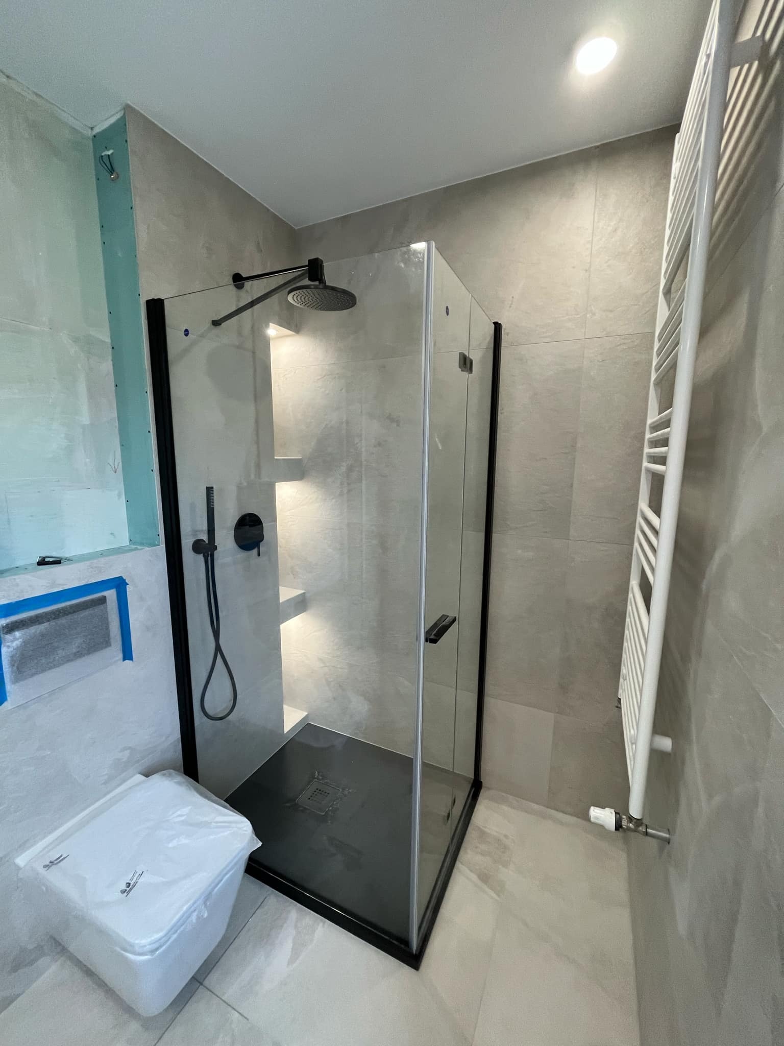 Wnętrze łazienki w trakcie remontu, z czarną kabiną prysznicową typu walk-in, białą drabinką grzejnikową i toaletą w folii ochronnej, widoczne niedokończone ściany z płyt karton-gips.