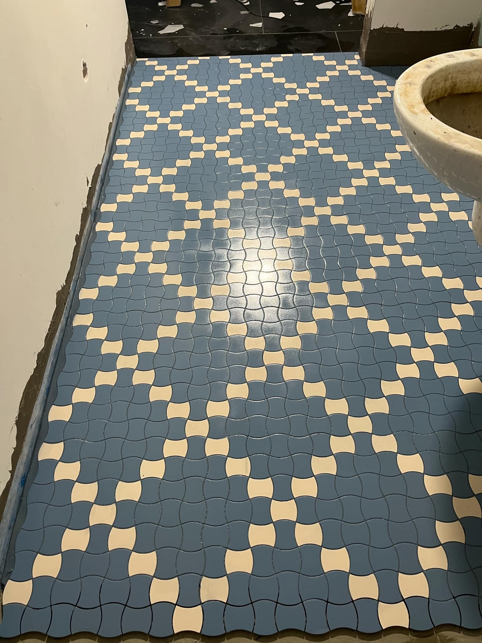 Niebiesko-biała mozaika podłogowa z zaokrąglonych płytek w łazience, widoczny fragment muszli klozetowej, ślady prac wykończeniowych przy ścianie.