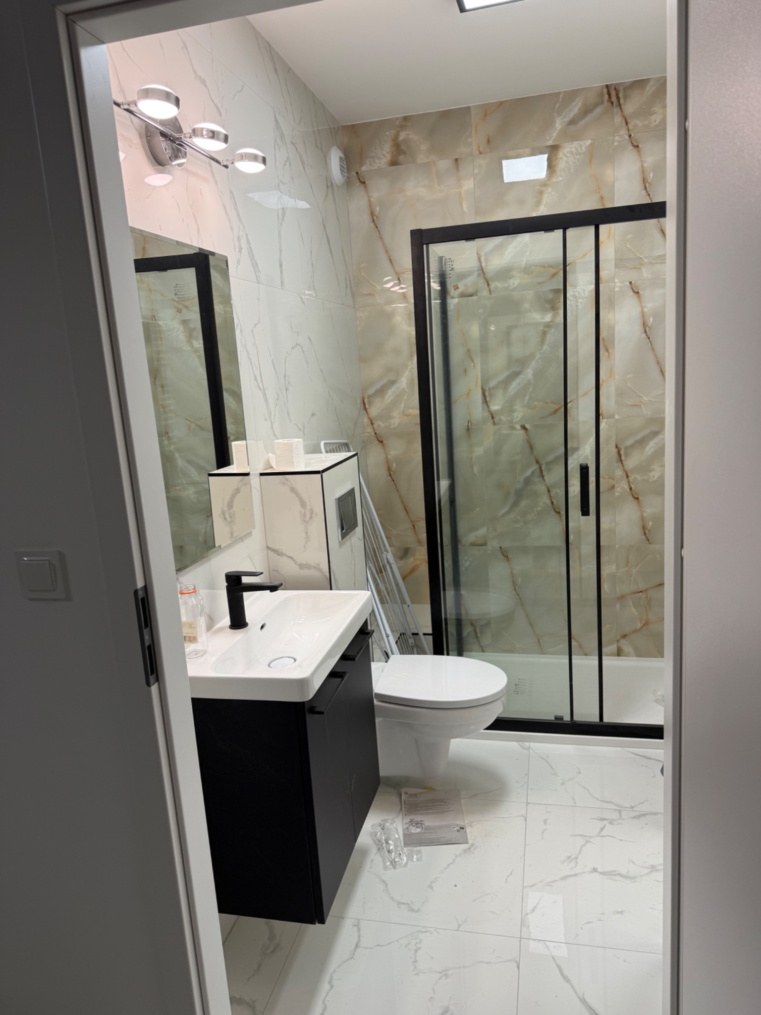 Nowoczesna łazienka z marmurowymi płytkami, czarną kabiną prysznicową i umywalką z szafką, widok zza drzwi, na podłodze resztki po remoncie.