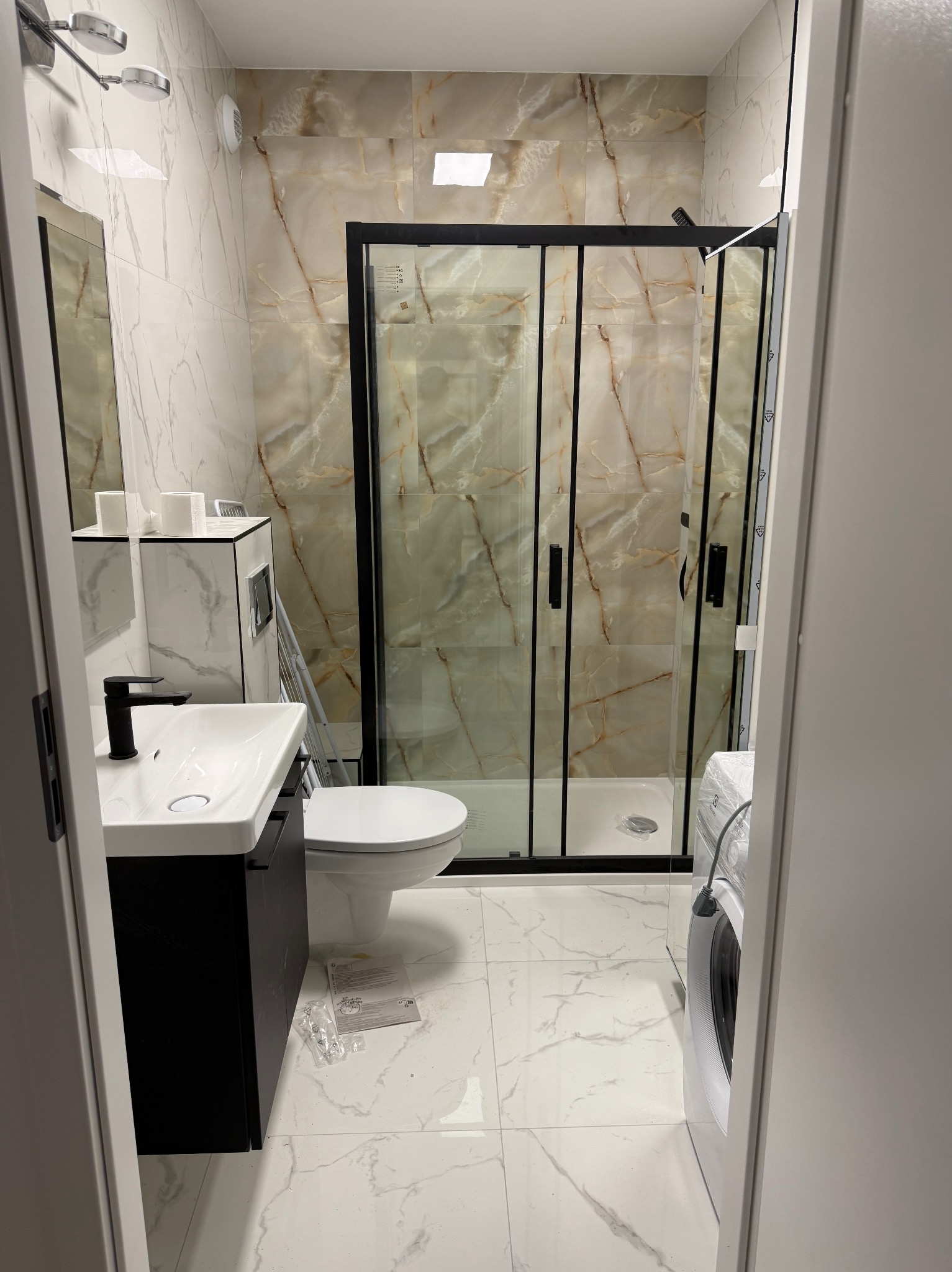 Nowoczesna łazienka w Warszawie z kabiną prysznicową z czarnymi ramami i marmurowymi płytkami, umywalka z czarną szafką, biała toaleta i pralka.