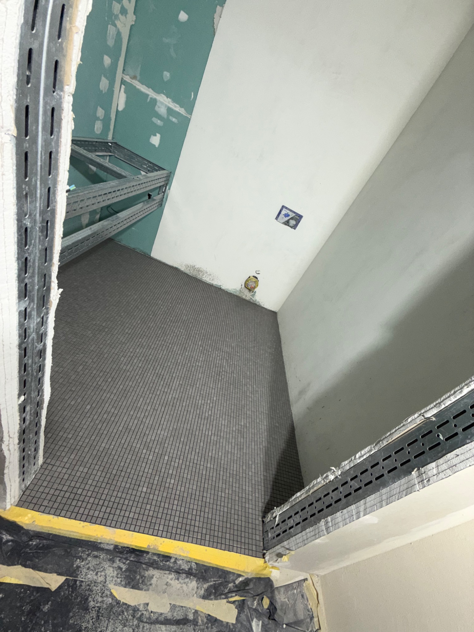 Wnętrze łazienki w trakcie prac wykończeniowych, widoczne szare mozaikowe płytki na podłodze, metalowe profile konstrukcyjne przy ścianach, częściowo wykończona ściana z płyty gipsowo-kartonowej...