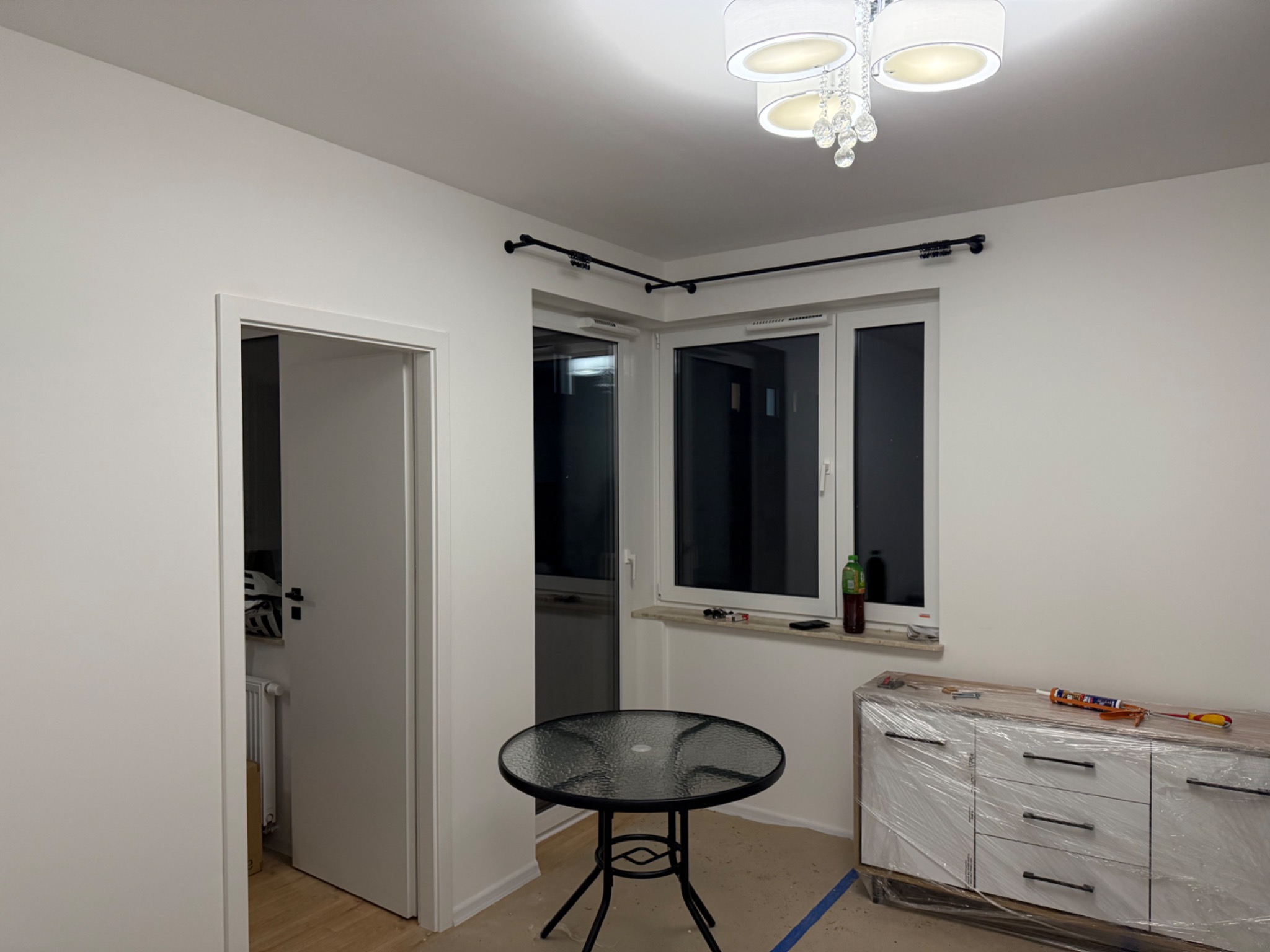 Wnętrze pokoju w trakcie prac wykończeniowych: widoczna listwa karniszowa zamontowana pod sufitem, okno, stół, komoda zabezpieczona folią i narzędzia na niej. Ściany pomalowane na biało.