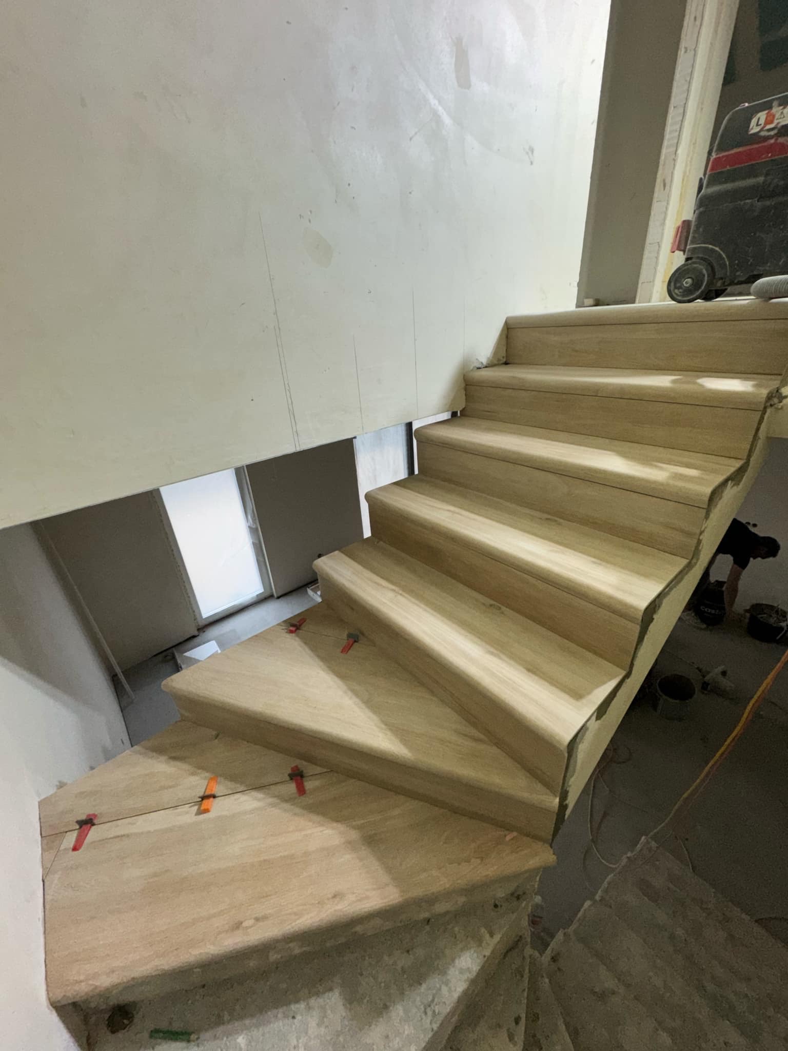 Montaż drewnopodobnych płytek schodowych z użyciem klipsów poziomujących, widoczny odkurzacz przemysłowy i surowe ściany w trakcie prac wykończeniowych.